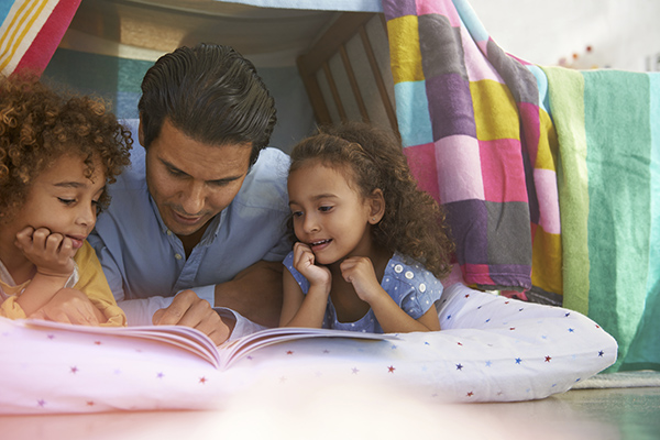 Shot of a young father lying in a tent fort with his children and reading them a book