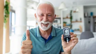 Older man smiles while holding a blood glucose meter and giving a thumbs-up at home.